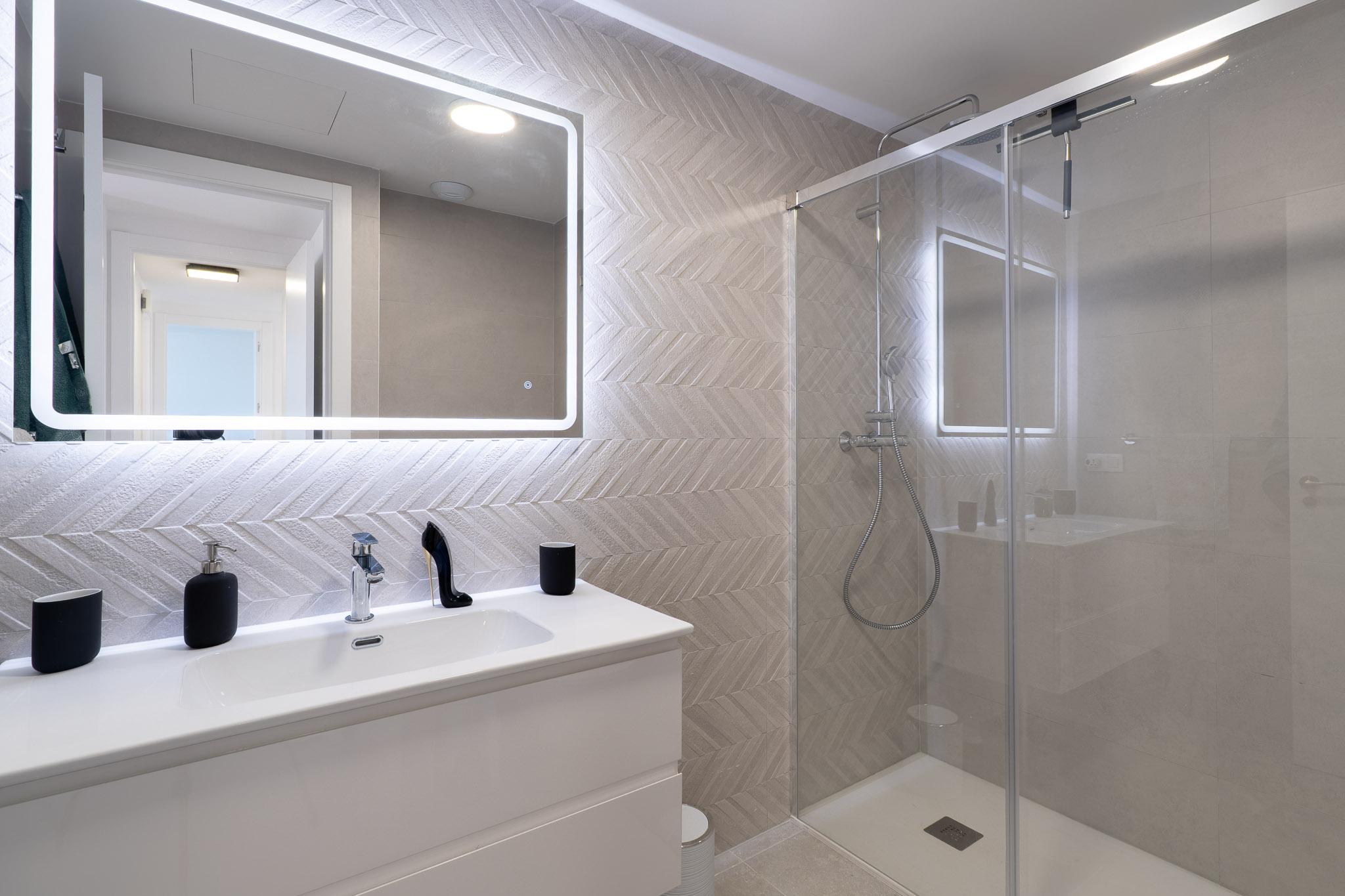 Modern bathroom featuring a white vanity, an illuminated mirror, and herringbone-patterned wall tiles. The glass-enclosed shower includes a rain shower and chrome fixtures.
