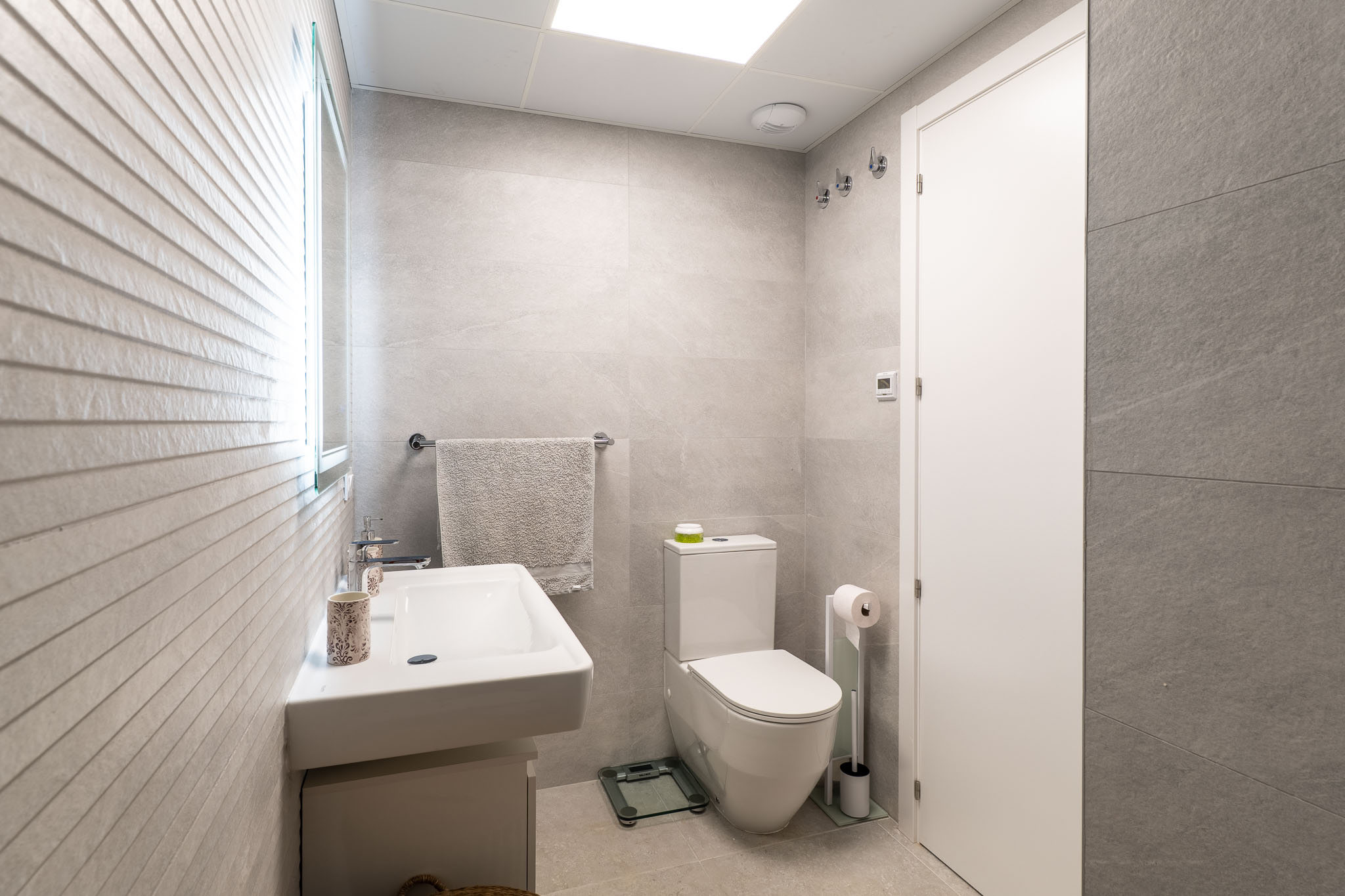 Modern bathroom featuring light grey large-format tiles, a wall-mounted sink, and a toilet. The illuminated mirror complements the textured accent wall.