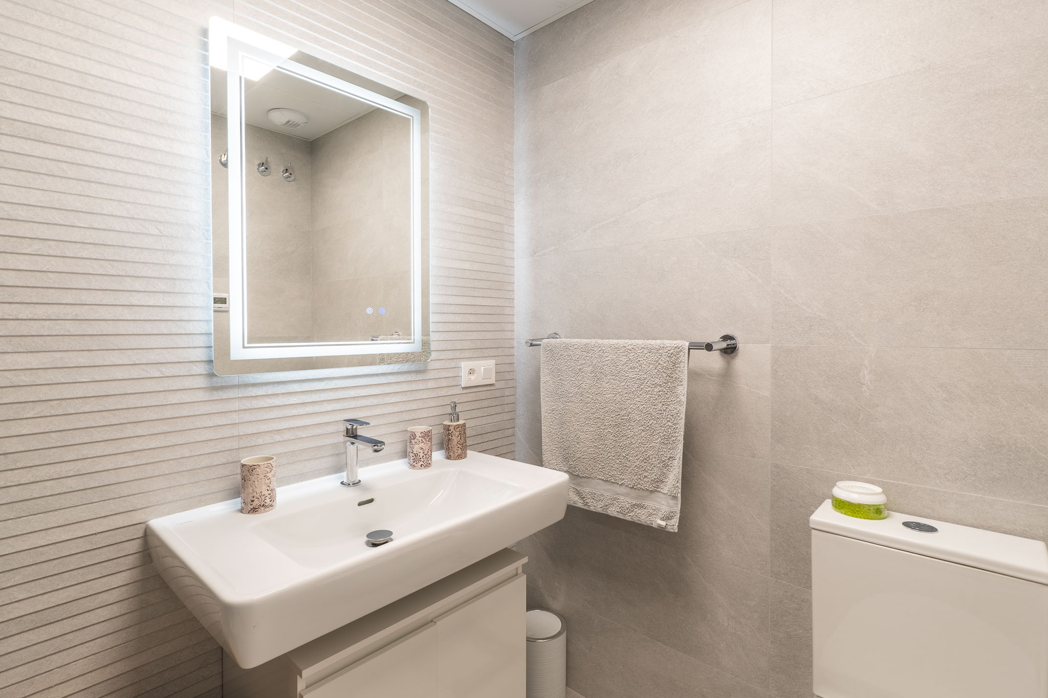 Modern bathroom vanity featuring a white rectangular sink with a wall-mounted faucet, an illuminated mirror, and light grey textured wall tiles.