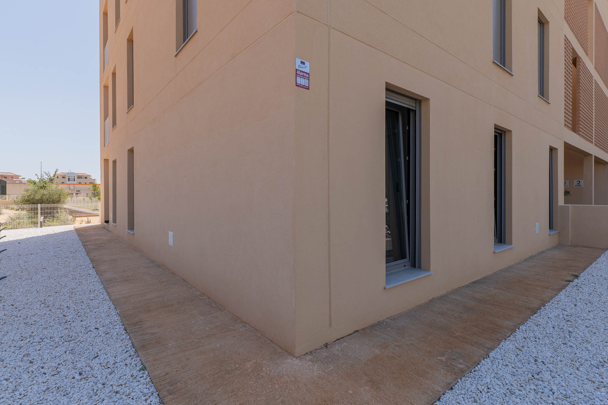 Modern building exterior featuring a light beige stucco facade with dark-framed windows. The ground level includes a textured concrete path alongside a white pebble landscape, with an alarm system visible.