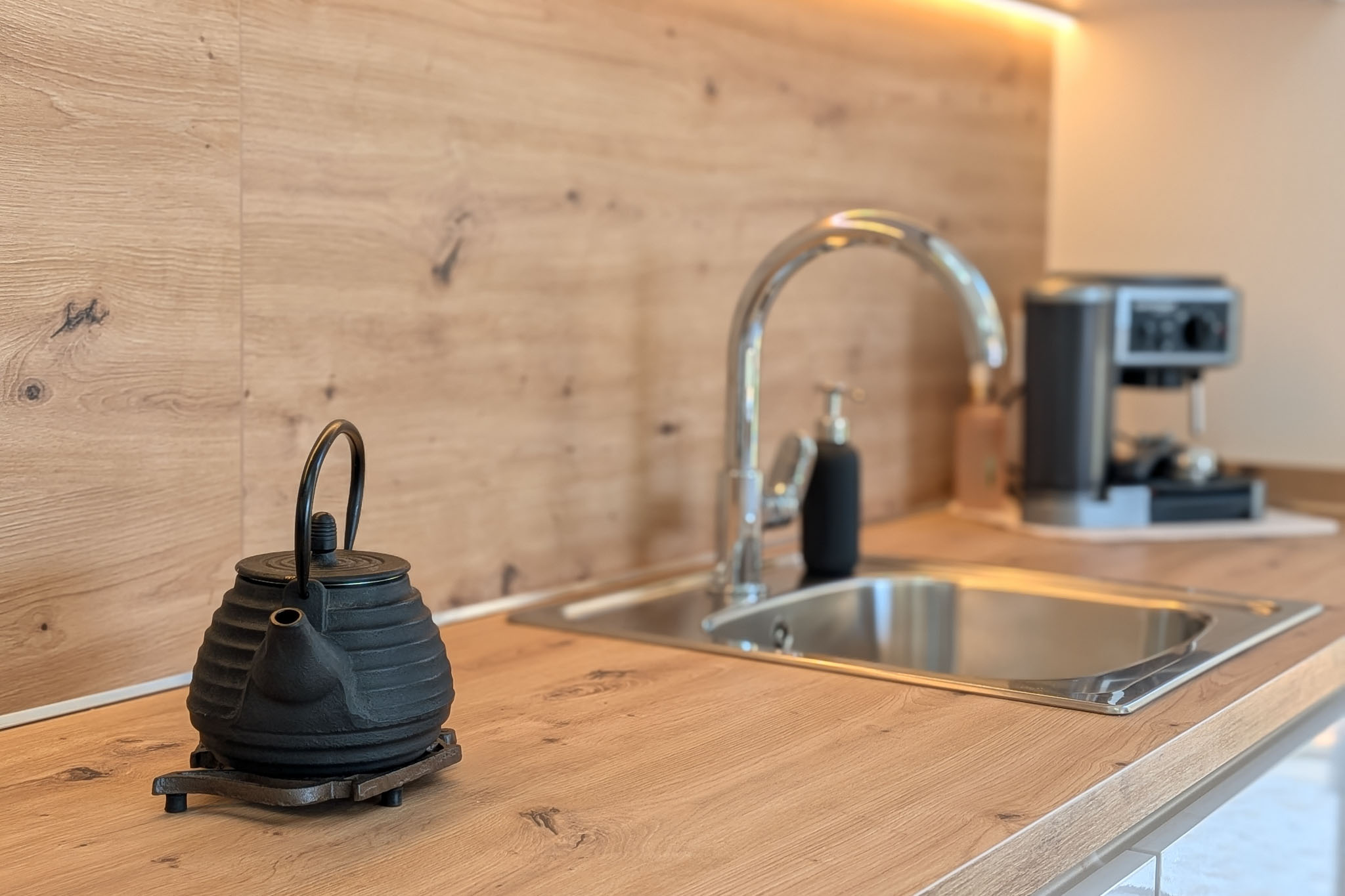 Modern kitchen countertop with light wood-look finish and matching backsplash. Features a stainless steel sink, chrome faucet, under-cabinet LED lighting, and a cast iron teapot in the foreground.