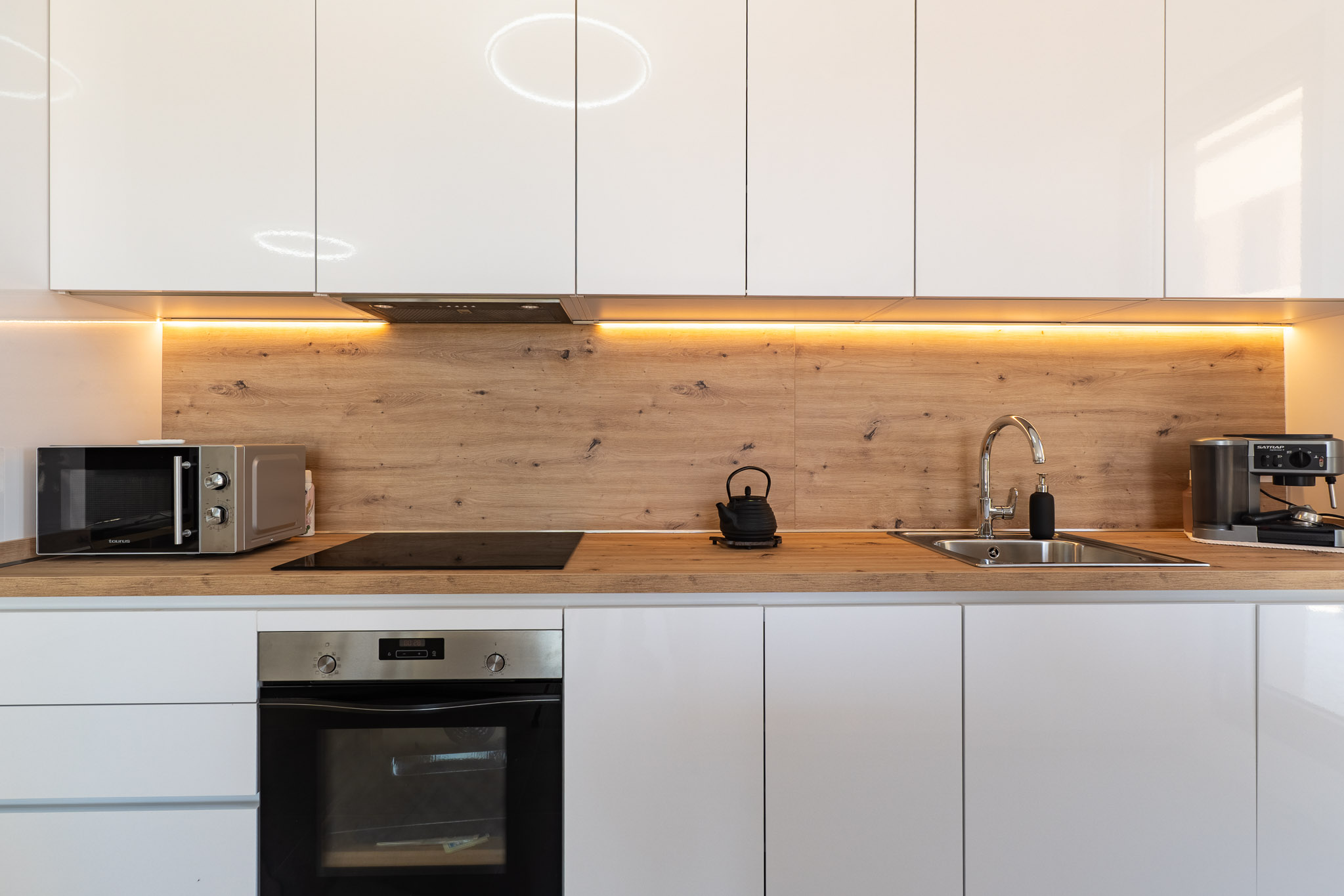 Modern kitchen featuring glossy white cabinetry, a wood-effect countertop and backsplash, and warm under-cabinet LED lighting. Includes integrated oven, induction hob, and microwave.