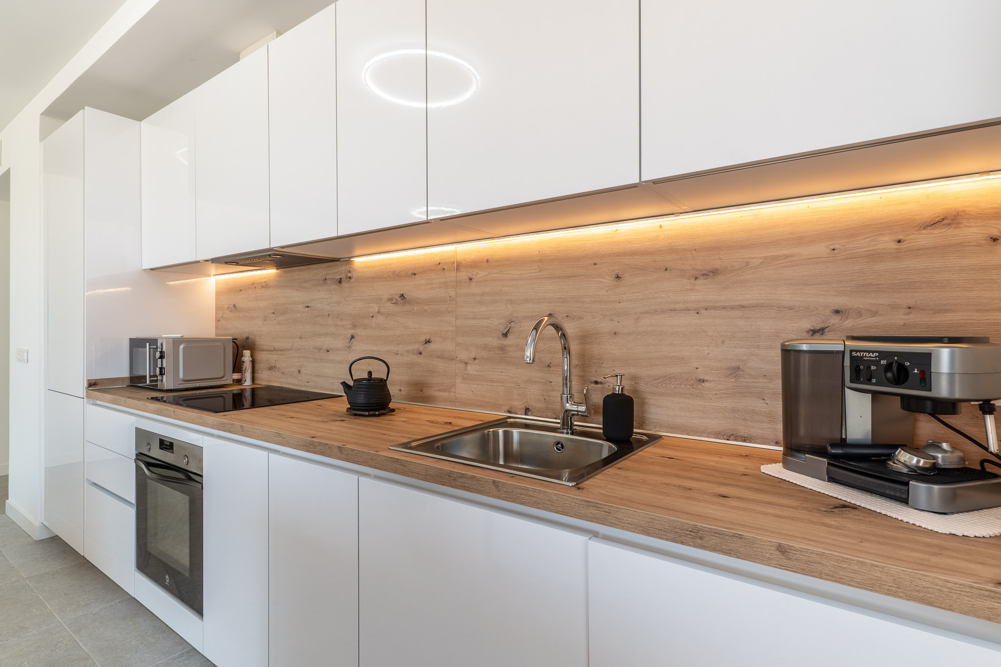 Modern kitchen featuring glossy white cabinetry, light wood-grain countertop, and matching backsplash. Equipped with an integrated oven, induction hob, and stainless steel sink, illuminated by under-cabinet LED lighting.