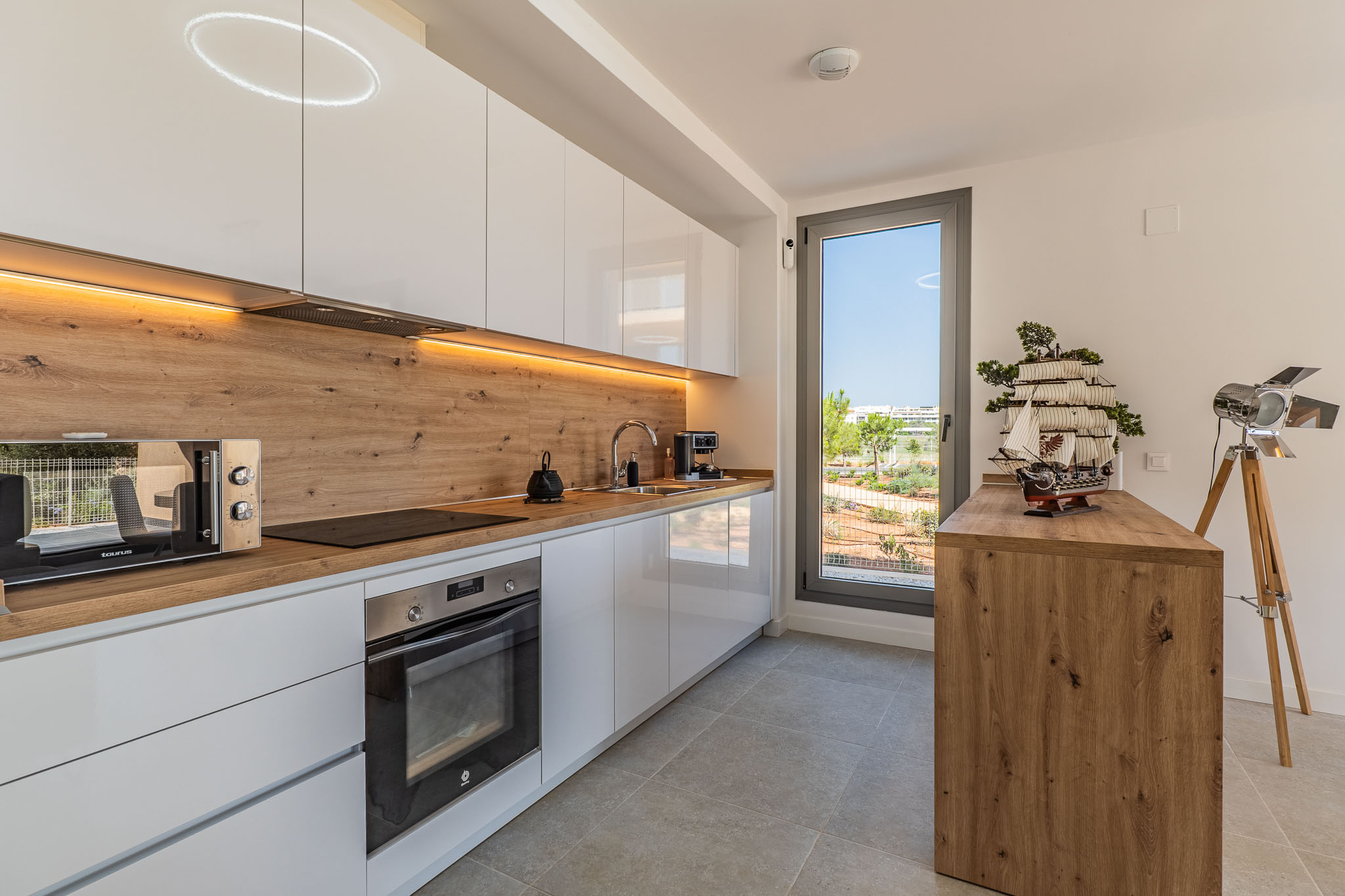 Modern kitchen featuring white high-gloss cabinets, wood-effect countertops, and integrated appliances. Under-cabinet LED lighting and a window offering views of the landscaped outdoor area.
