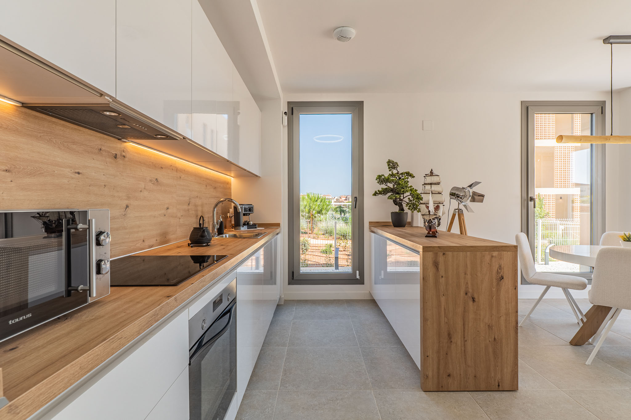 Modern open-plan kitchen featuring glossy white cabinetry, wood-effect countertops and backsplash, with integrated appliances and under-cabinet LED lighting. Offers direct access to the outdoor area.