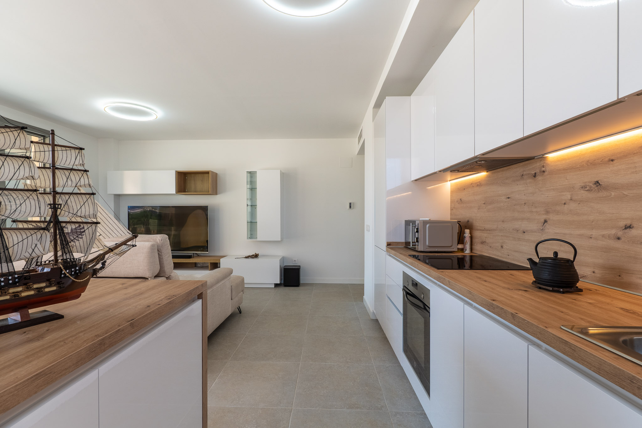 Open-plan living and dining area featuring a modern kitchen with white high-gloss cabinets, light wood-effect countertops, integrated appliances, and warm LED under-cabinet lighting. Grey tiled flooring unifies the space.
