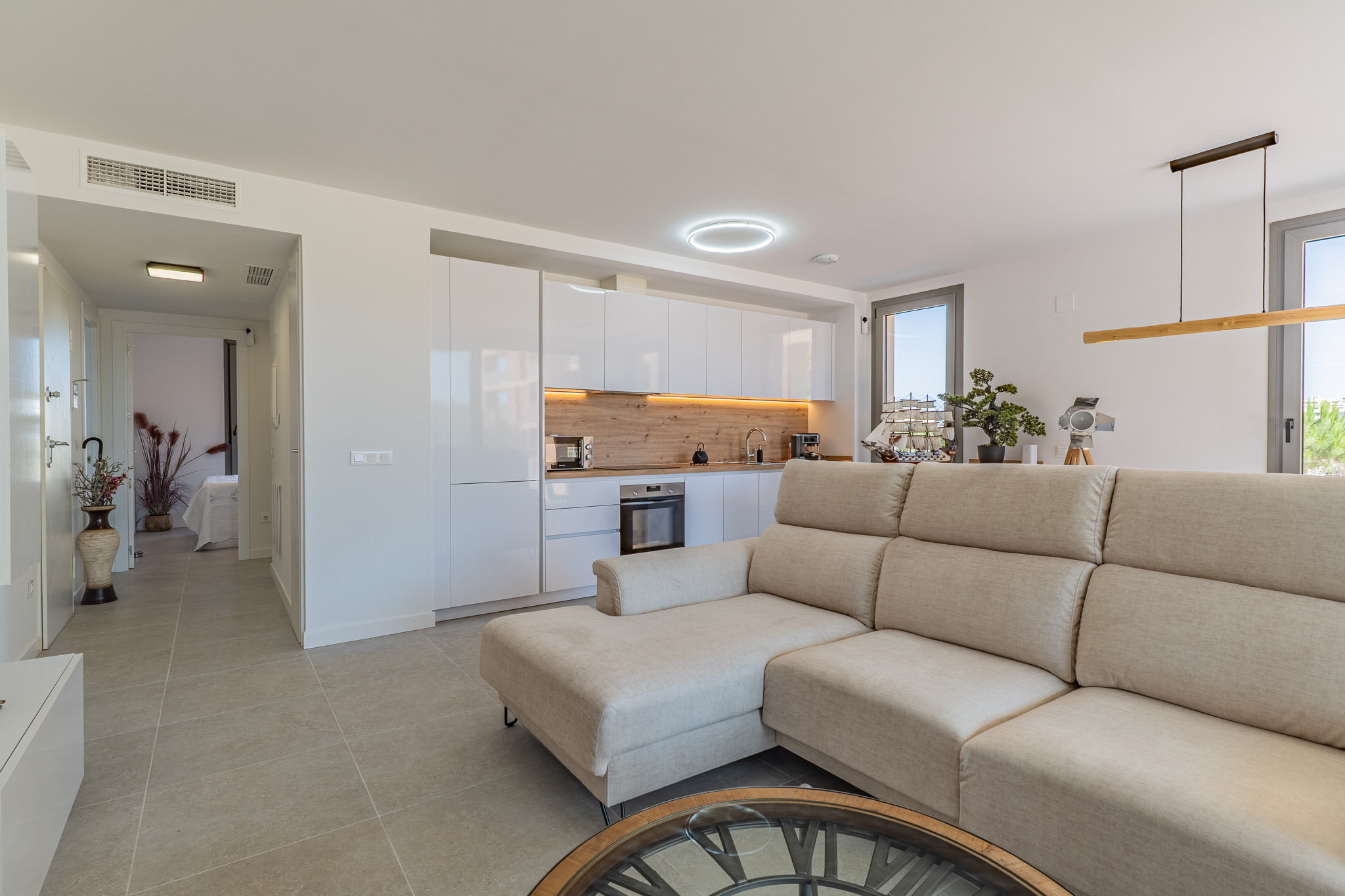 Open-plan living room and kitchen featuring a beige sectional sofa, modern white cabinetry, and a light wood backsplash. Tiled floor and natural light from large windows.
