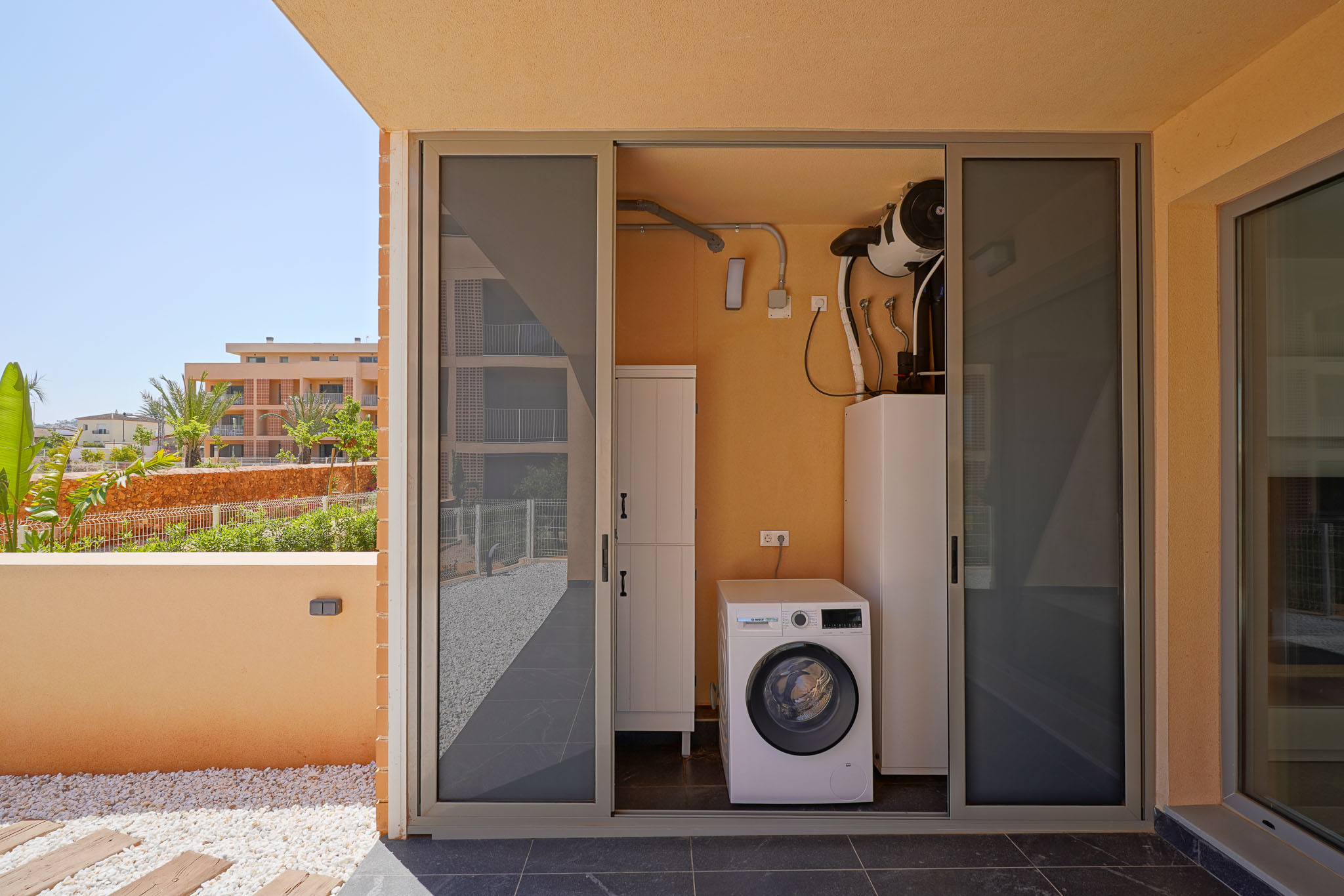Outdoor utility room featuring a Bosch washing machine and aerothermal system, accessed from a tiled terrace with decorative pebbles and garden views.