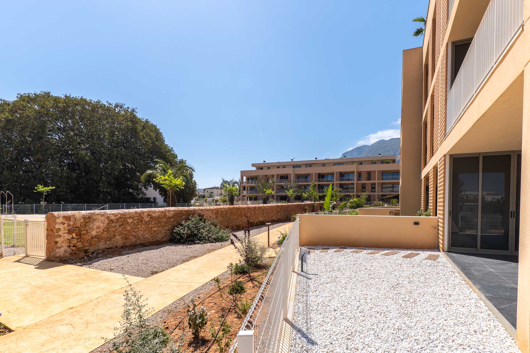 Private ground-floor garden with white gravel and stepping stones, bordered by a low white fence and a rustic stone wall. Views of modern buildings and distant mountains.