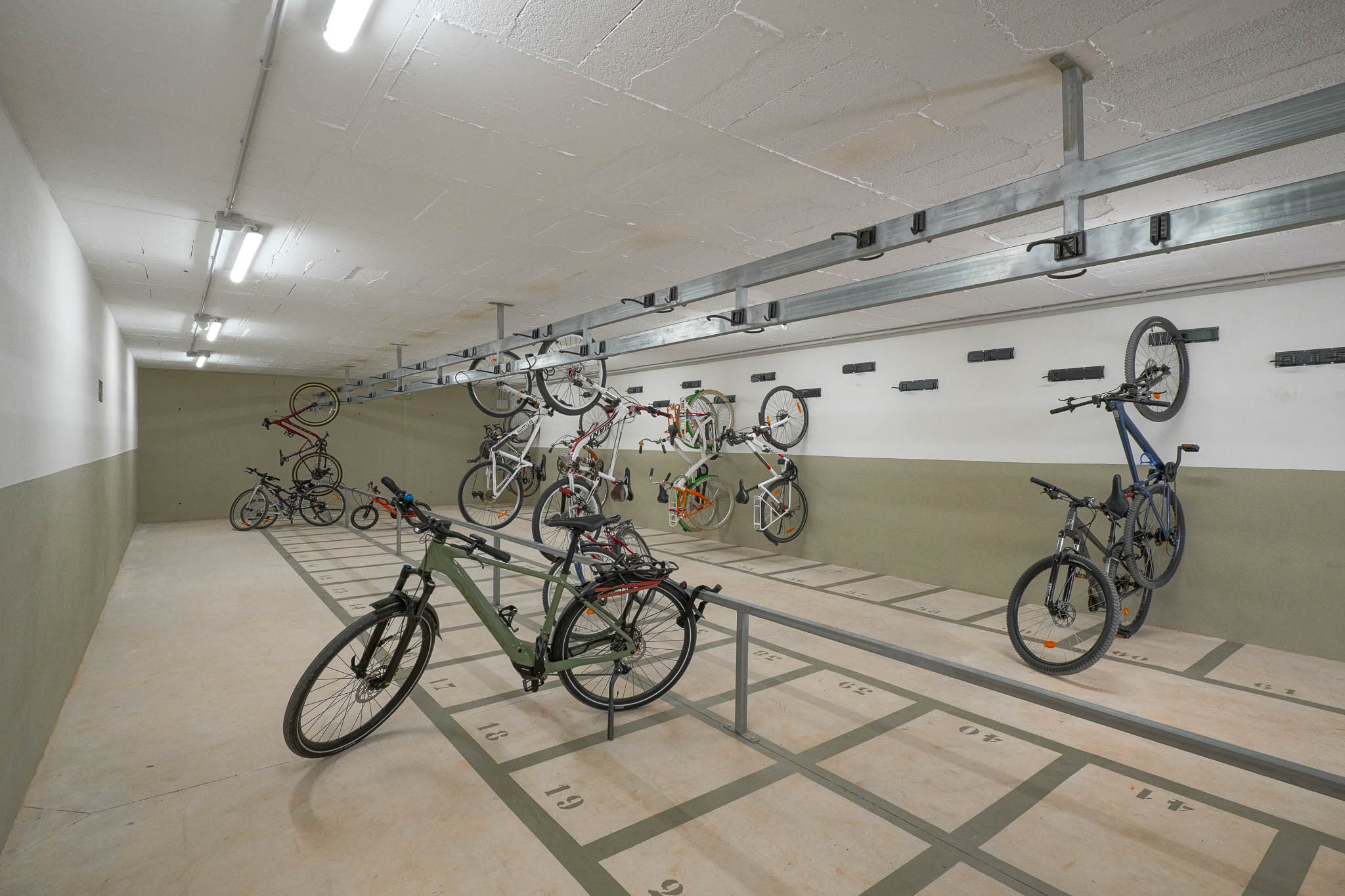 Spacious communal bicycle parking area featuring numbered floor spaces and wall-mounted racks. Brightly lit by fluorescent ceiling lights, offering secure storage within the residential complex.