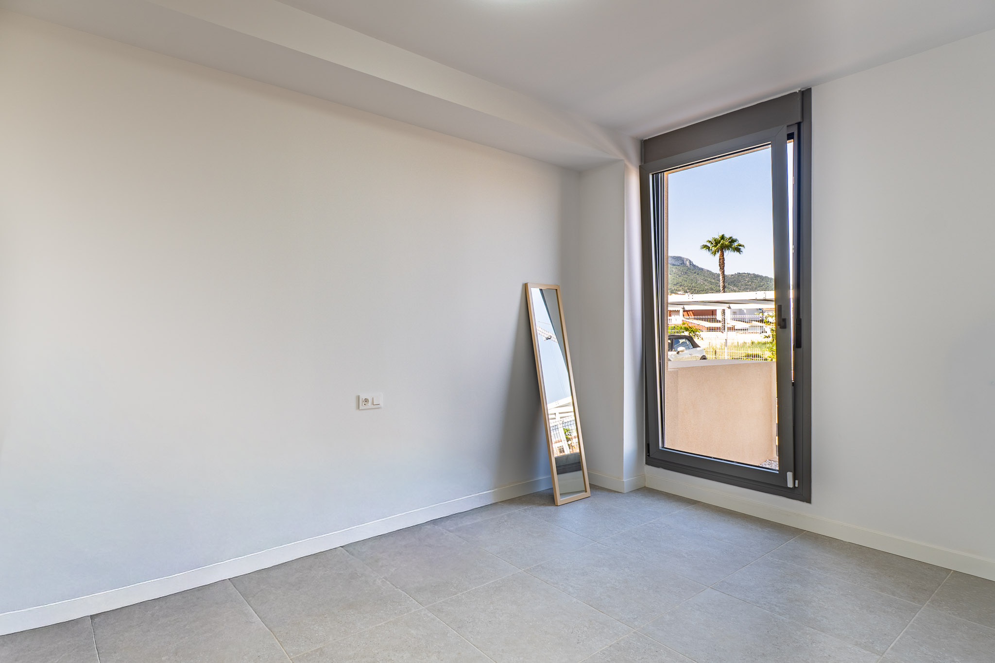 Unfurnished room featuring white walls and grey tiled flooring, ready for customization. Large window offers natural light and views of a palm tree and distant mountains.