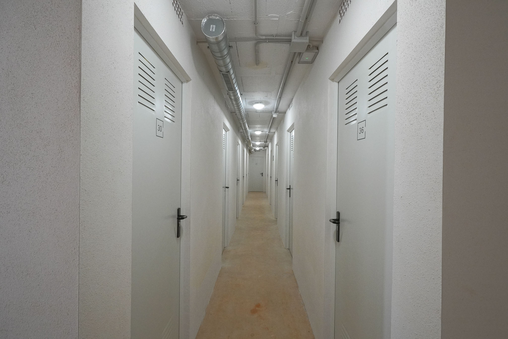 Utilitarian corridor leading to individual storage units, featuring numbered white doors with ventilation slats. Exposed concrete ceiling displays industrial ducting and recessed lighting, complementing textured walls.