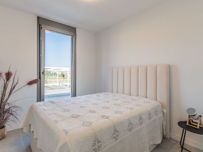 Bright bedroom featuring a bed with a tall, padded beige headboard. A large floor-to-ceiling window offers exterior views. Grey tiled floor and a decorative plant complete the space.