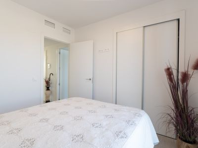 Bright bedroom featuring a bed with a white patterned quilt. Equipped with a built-in sliding wardrobe and integrated ceiling air conditioning vents. A decorative plant adds a touch of color.