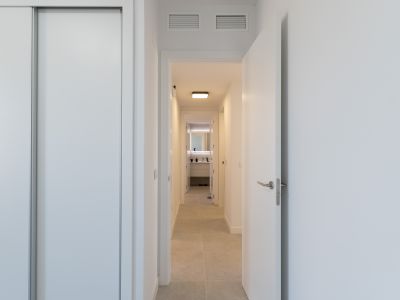 Bright hallway featuring a white sliding wardrobe and an open door. Light-toned floor tiles lead to a modern bathroom with a backlit mirror and contemporary vanity. Ceiling grilles visible.