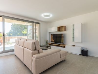 Bright living room featuring a beige sectional sofa, light grey tiled floor, and large sliding glass doors leading to the outdoor area. Modern media console with white and wood shelving.