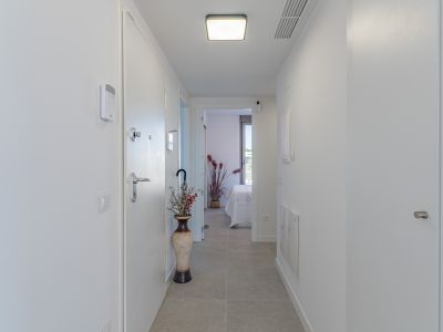 Bright, modern hallway with light grey tiled flooring and white walls. Features include a security panel, a contemporary ceiling light, and a glimpse into a bedroom.