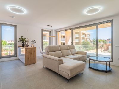 Bright open-plan living-dining area with a beige sectional sofa and oval coffee table. Large glass doors provide abundant natural light and residential views. Features an open kitchen with a wooden counter.