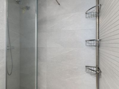 Contemporary shower area with large format light grey tiles, a textured accent wall, clear glass screen, and a practical chrome corner storage unit.