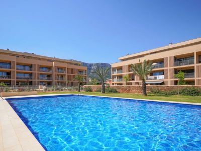 Expansive communal swimming pool with blue mosaic tiles, bordered by green lawn and modern residential buildings featuring balconies. Palm trees and mountains are visible in the background.