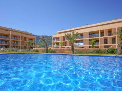 Expansive communal swimming pool with shimmering blue mosaic tiles, set against modern residential buildings featuring balconies and a distant mountain range under a clear sky.