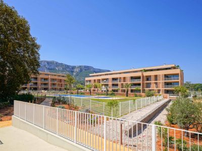 Exterior view of the modern residential complex featuring two light-colored buildings, a communal swimming pool, and landscaped gardens. Mountains are visible in the background.