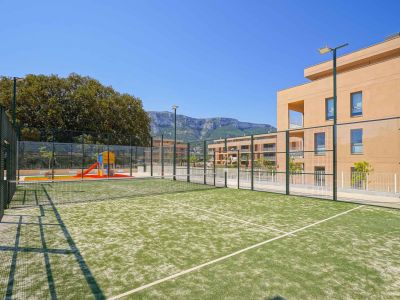 Green artificial turf padel court, fenced and equipped with lighting poles, adjacent to a children's playground and modern residential buildings with mountain views.