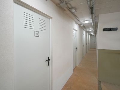 Interior corridor featuring a series of white storage unit doors with ventilation slats and black handles. Exposed ceiling ducts and recessed lighting illuminate the concrete floor.