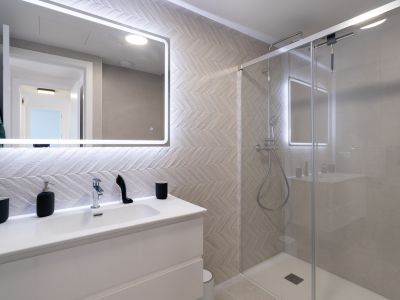 Modern bathroom featuring a white vanity, an illuminated mirror, and herringbone-patterned wall tiles. The glass-enclosed shower includes a rain shower and chrome fixtures.
