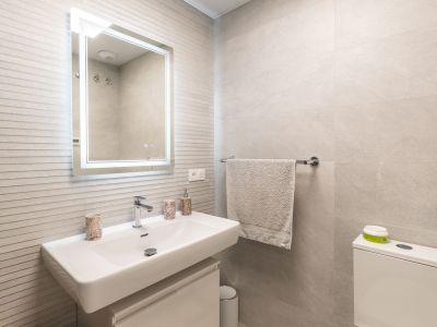 Modern bathroom vanity featuring a white rectangular sink with a wall-mounted faucet, an illuminated mirror, and light grey textured wall tiles.