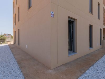 Modern building exterior featuring a light beige stucco facade with dark-framed windows. The ground level includes a textured concrete path alongside a white pebble landscape, with an alarm system visible.
