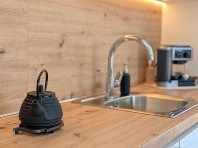 Modern kitchen countertop with light wood-look finish and matching backsplash. Features a stainless steel sink, chrome faucet, under-cabinet LED lighting, and a cast iron teapot in the foreground.