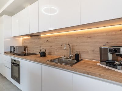 Modern kitchen featuring glossy white cabinetry, light wood-grain countertop, and matching backsplash. Equipped with an integrated oven, induction hob, and stainless steel sink, illuminated by under-cabinet LED lighting.