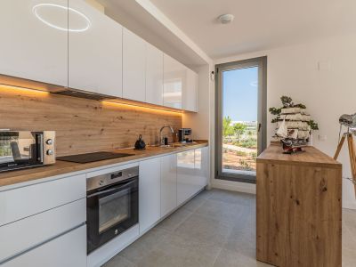 Modern kitchen featuring white high-gloss cabinets, wood-effect countertops, and integrated appliances. Under-cabinet LED lighting and a window offering views of the landscaped outdoor area.