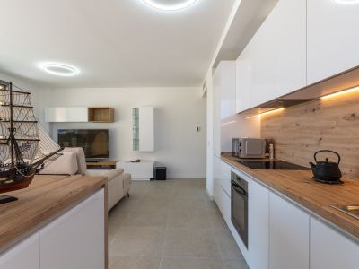 Open-plan living and dining area featuring a modern kitchen with white high-gloss cabinets, light wood-effect countertops, integrated appliances, and warm LED under-cabinet lighting. Grey tiled flooring unifies the space.
