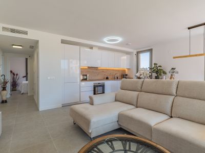 Open-plan living room and kitchen featuring a beige sectional sofa, modern white cabinetry, and a light wood backsplash. Tiled floor and natural light from large windows.