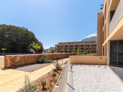 Private ground-floor garden with white gravel and stepping stones, bordered by a low white fence and a rustic stone wall. Views of modern buildings and distant mountains.