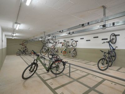 Spacious communal bicycle parking area featuring numbered floor spaces and wall-mounted racks. Brightly lit by fluorescent ceiling lights, offering secure storage within the residential complex.