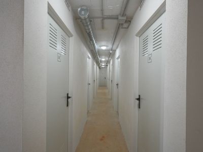 Utilitarian corridor leading to individual storage units, featuring numbered white doors with ventilation slats. Exposed concrete ceiling displays industrial ducting and recessed lighting, complementing textured walls.