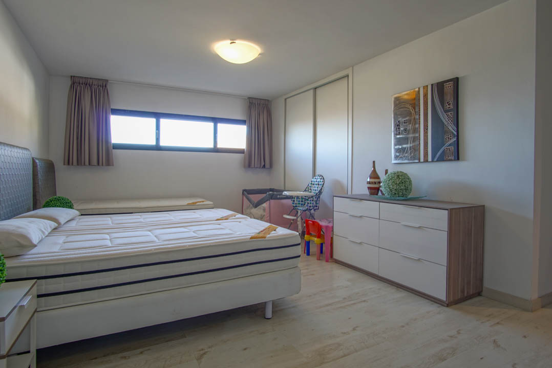 A bright bedroom featuring a bed with two mattresses, light wood-look flooring, and a long horizontal window. The room includes a white sliding wardrobe and a modern dresser with an abstract artwork above.