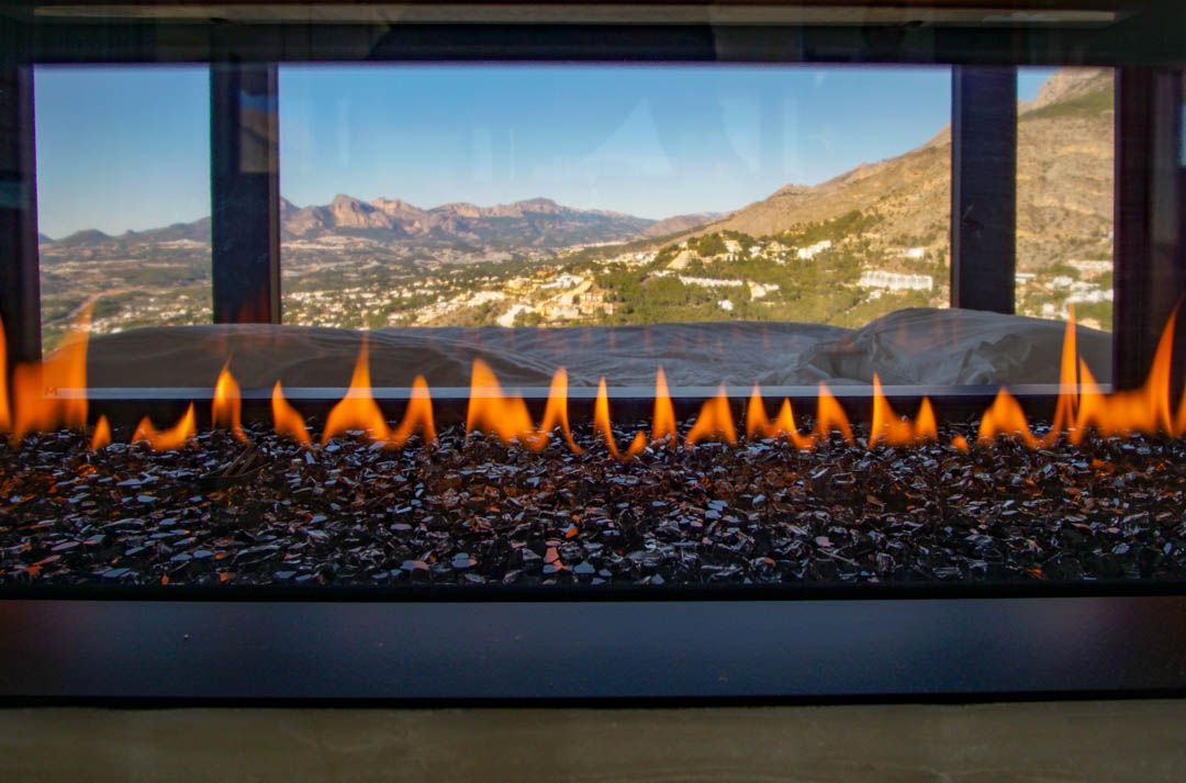 A modern gas fireplace with active flames over dark glass media, set against a panoramic window revealing distant mountains and a valley landscape.