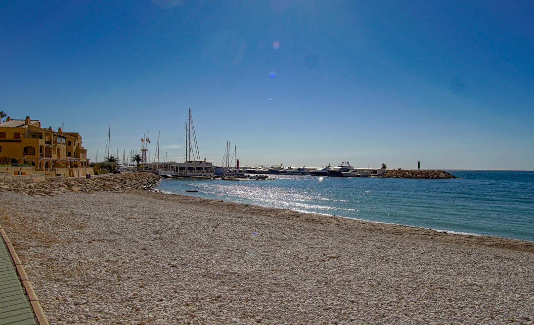 A wide pebble beach stretches towards a marina filled with yachts, flanked by traditional coastal buildings under a bright, clear blue sky.