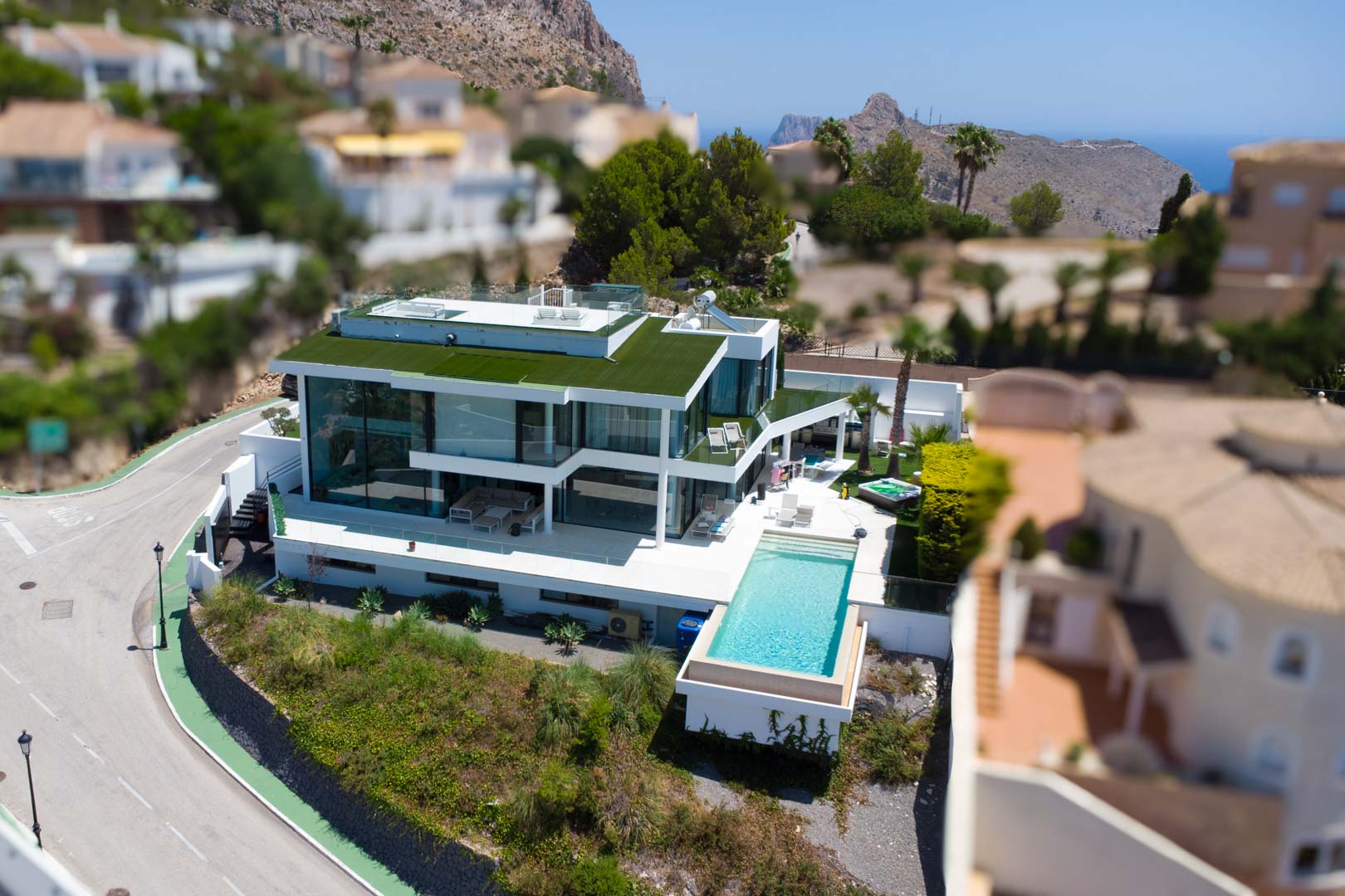Aerial view of a contemporary white villa with an infinity pool, extensive glass facades, a green roof, and a separate jacuzzi, offering mountain and sea views.