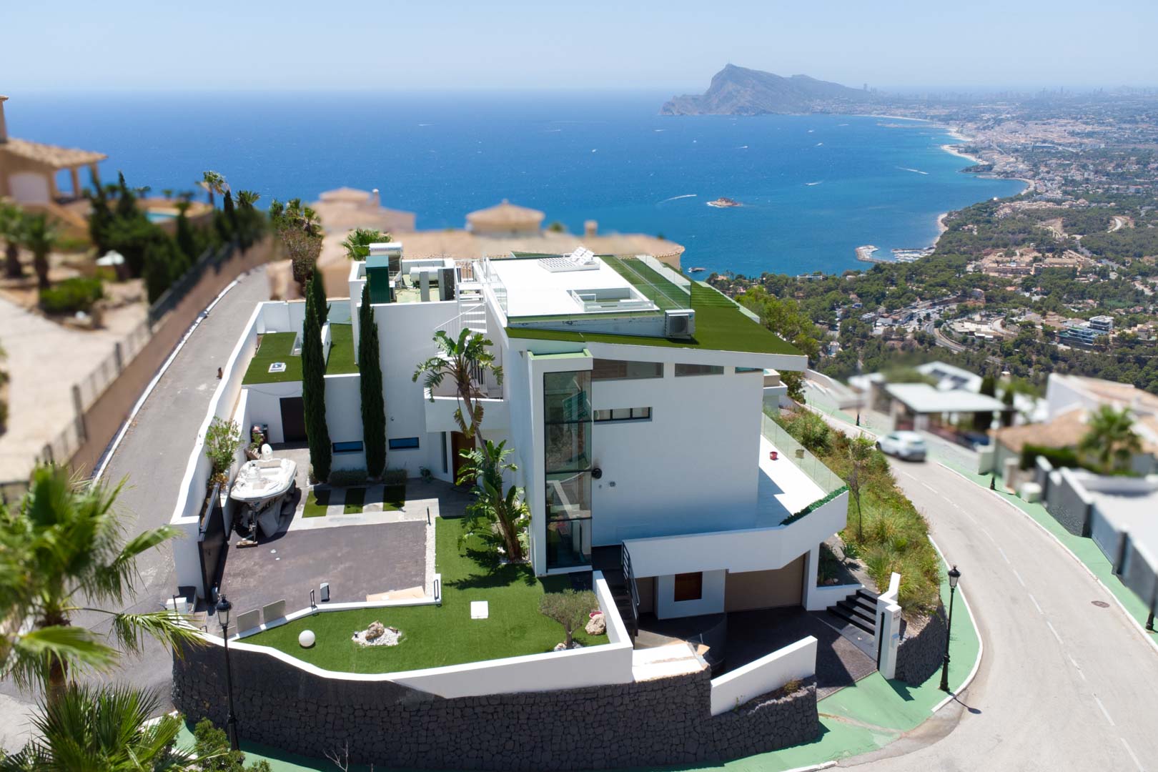 Aerial view of a modern white villa featuring multi-level architecture, green flat roofs, and an external glass elevator. The property includes a private driveway and offers panoramic sea and coastal views.
