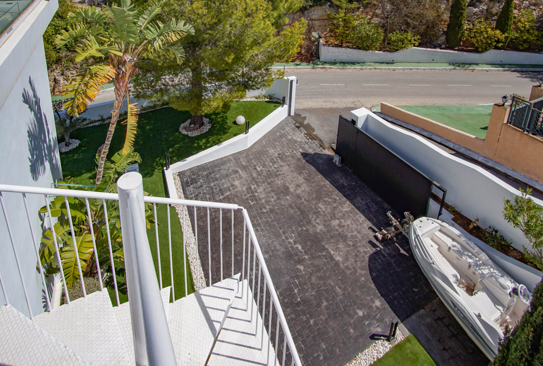 Aerial view of the main entrance featuring a white spiral staircase, a dark paved driveway, and artificial grass areas with a palm tree. An automatic gate and parking space are visible.