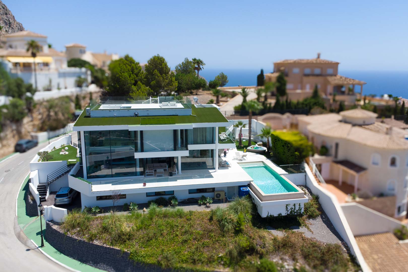 Aerial view of the modern white designer villa featuring a green roof, infinity pool, and separate jacuzzi, set against a sea view backdrop.
