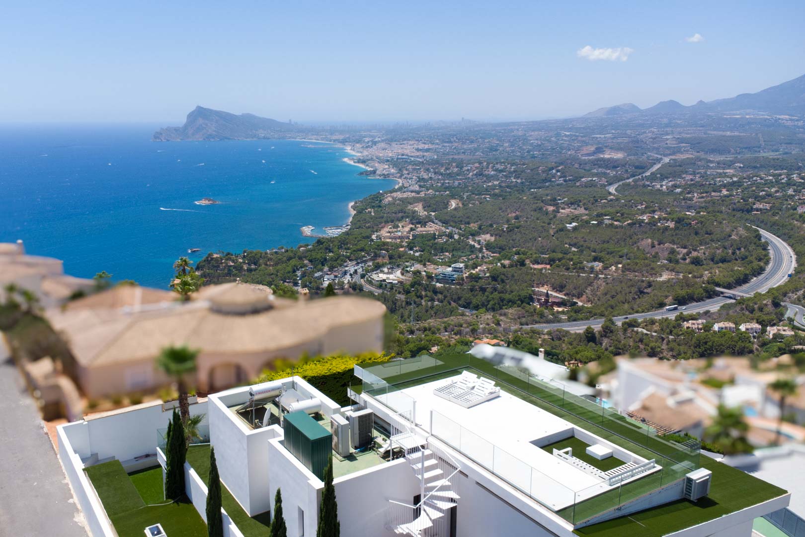 Aerial view of the villa's rooftop terrace featuring a white spiral staircase, artificial grass, glass railings, and expansive views of the Mediterranean Sea and coastal landscape.