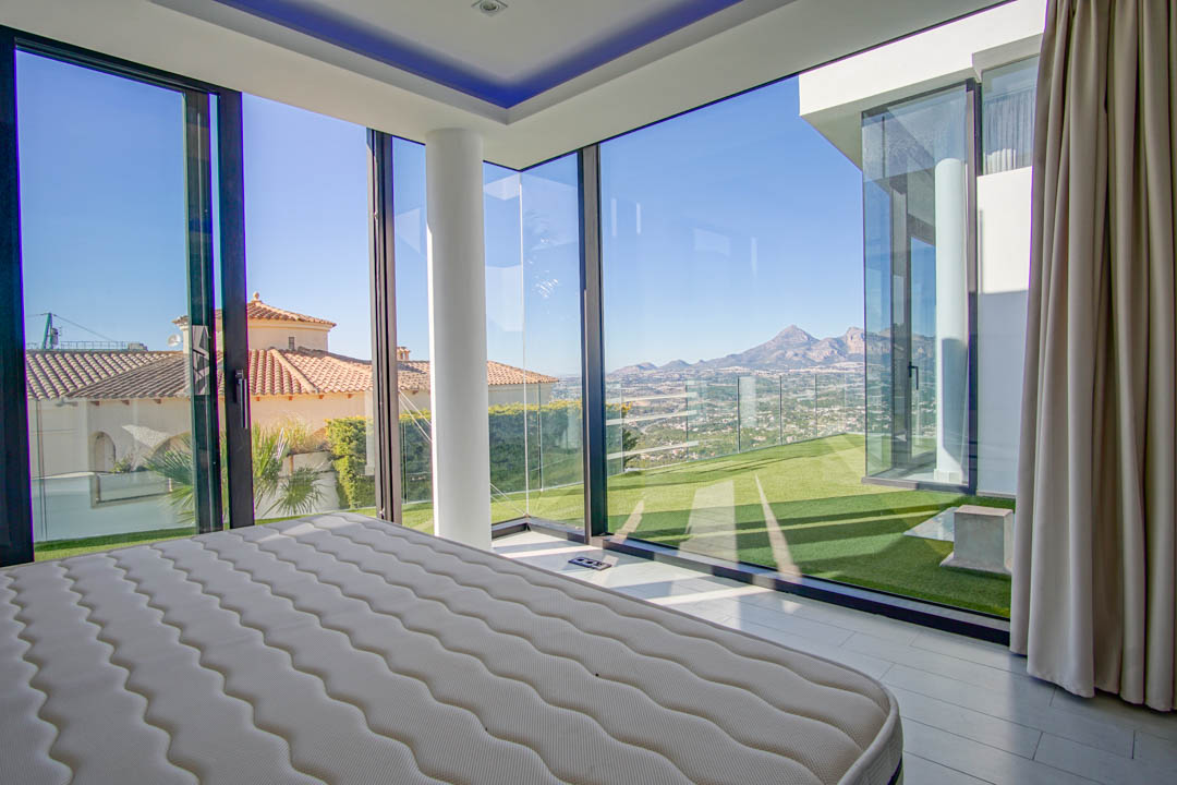 Bedroom featuring expansive corner glass walls, offering panoramic views of distant mountains, a valley, and neighboring rooftops. Includes a white structural pillar and direct access to an artificial lawn.