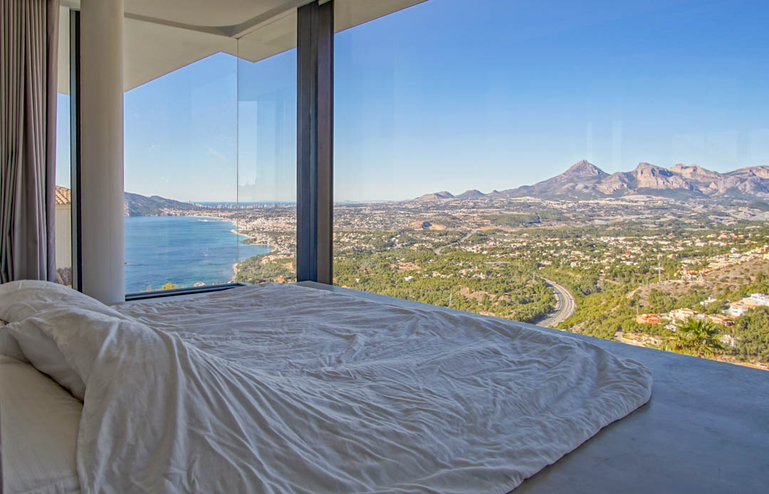 Bedroom featuring floor-to-ceiling glass, framing panoramic views of the Mediterranean Sea, a coastal town, and distant mountains. A low-profile bed with white linens faces the expansive vista.