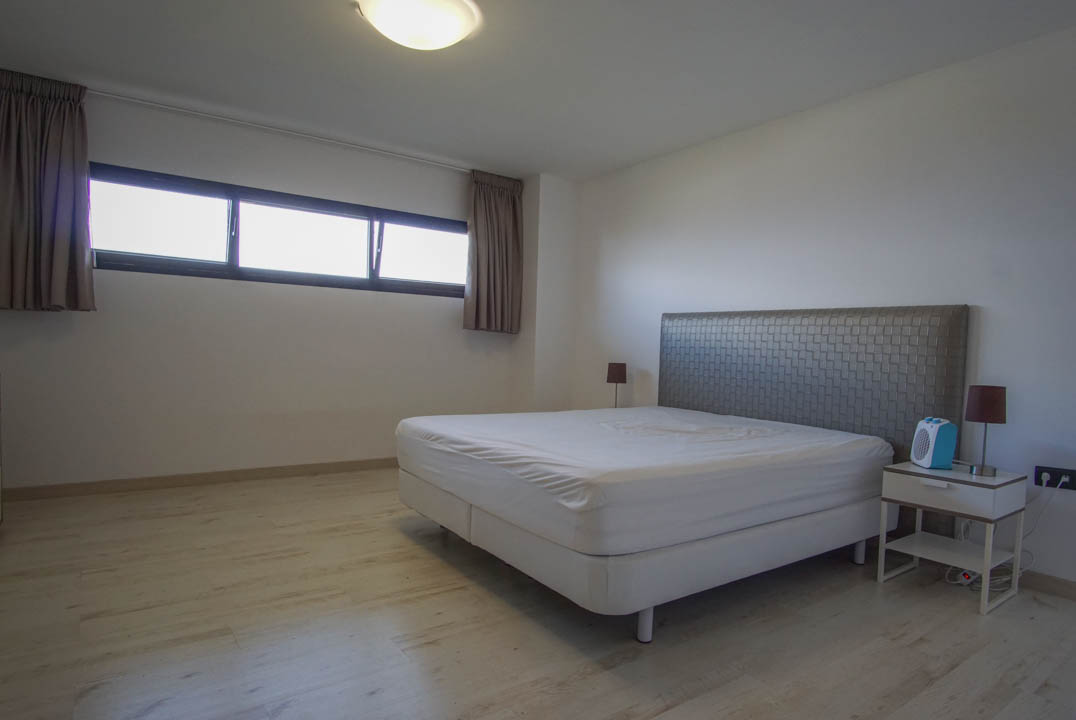 Bedroom featuring light wood-look flooring, white walls, and a bed with a textured grey headboard. A horizontal window with dark frames and curtains provides natural light.